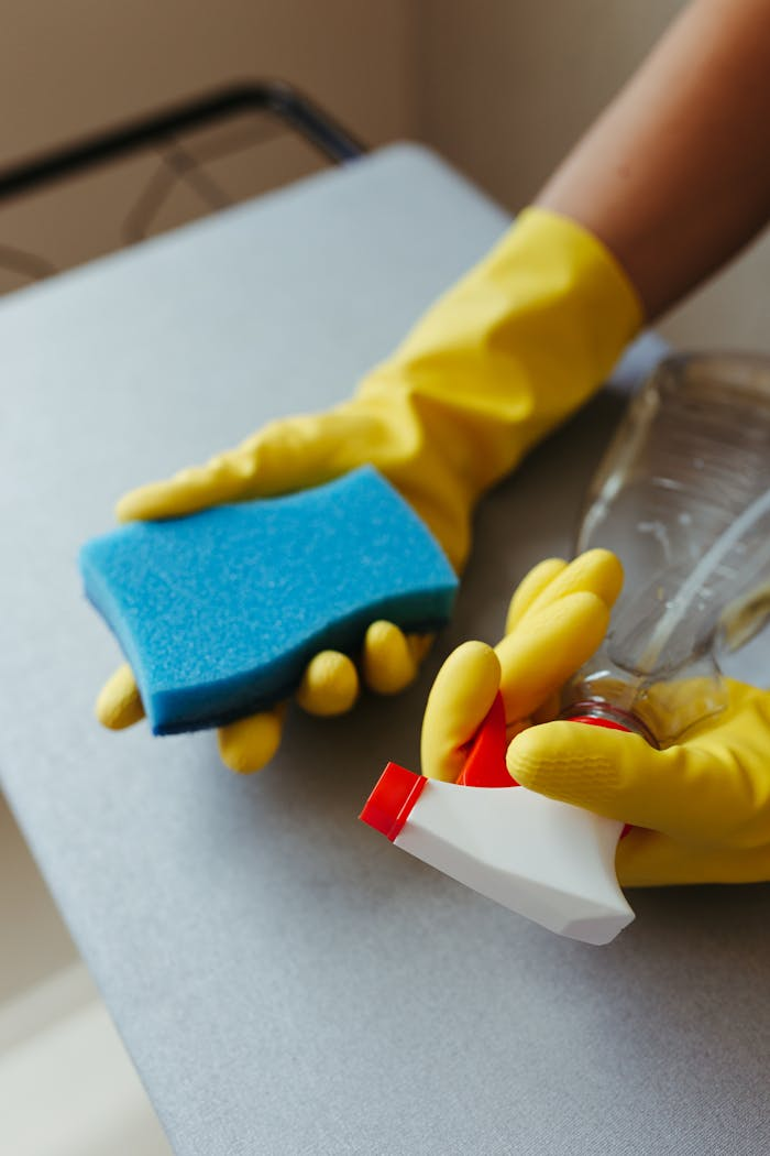 Close-up of a person cleaning a surface with a blue sponge and spray bottle, using yellow gloves.