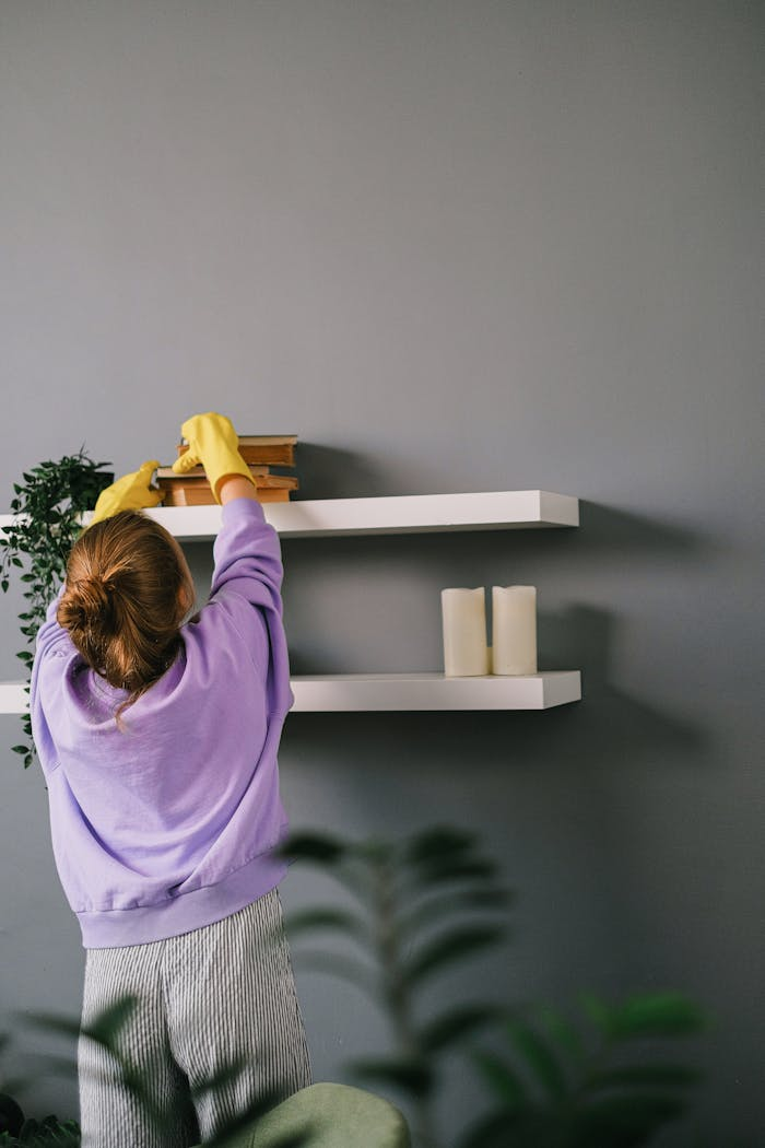 Back view of unrecognizable female in casual clothes and latex gloves standing and taking books while cleaning shelves in room with gray wall
