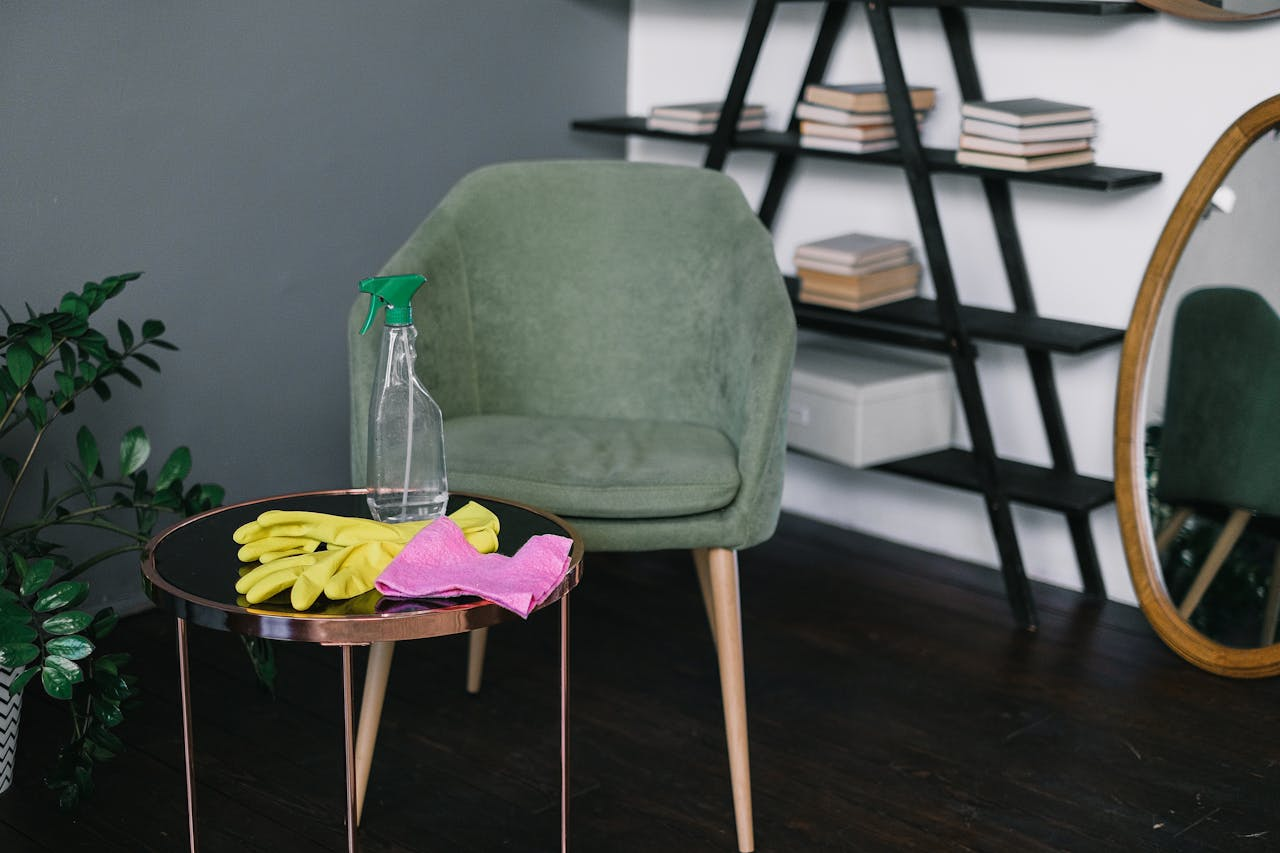 Yellow latex gloves placed near spray detergent bottle and rag on round table against comfortable chair in living room in daytime